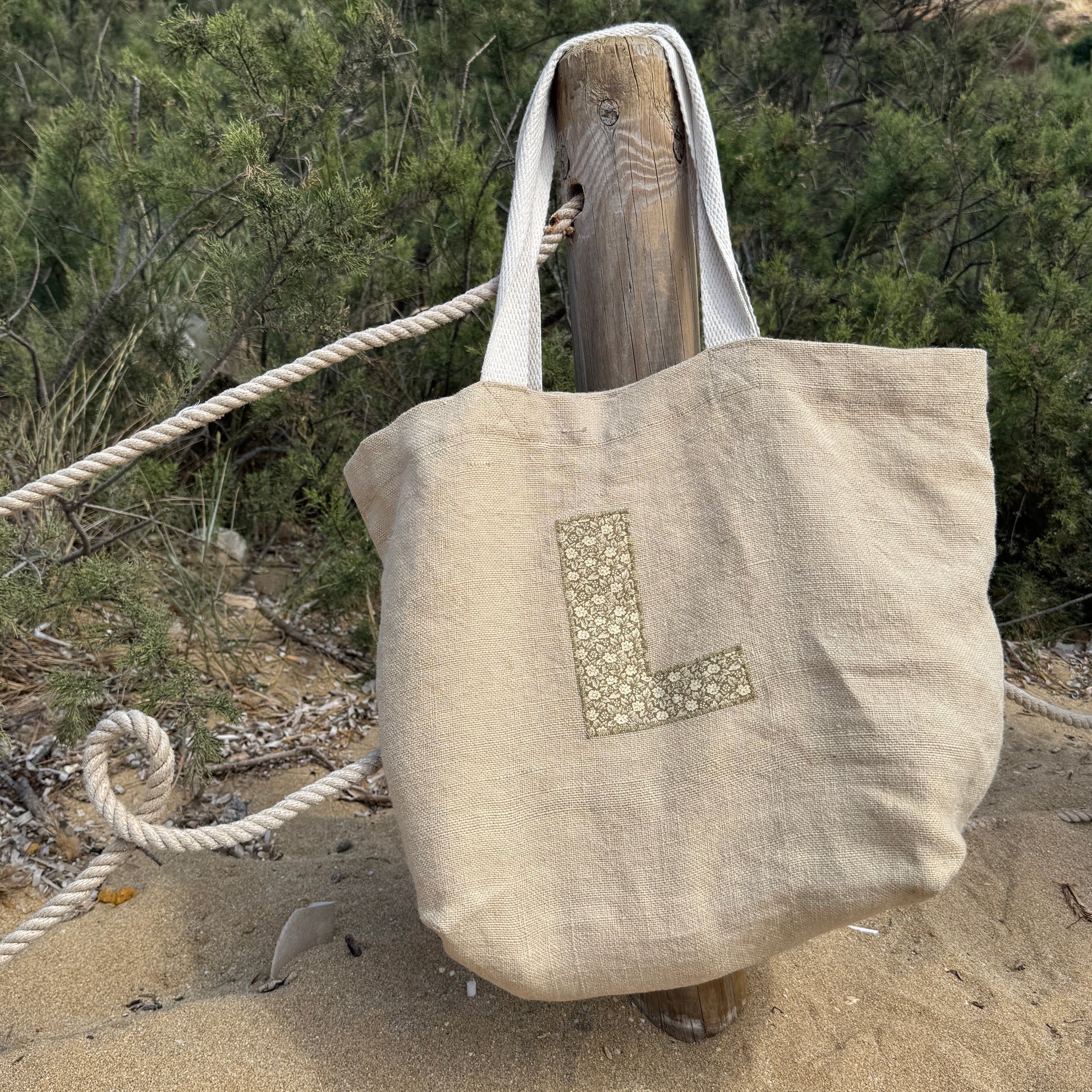 Beige tote bag with a letter 'L' on a wooden post in a natural beach setting