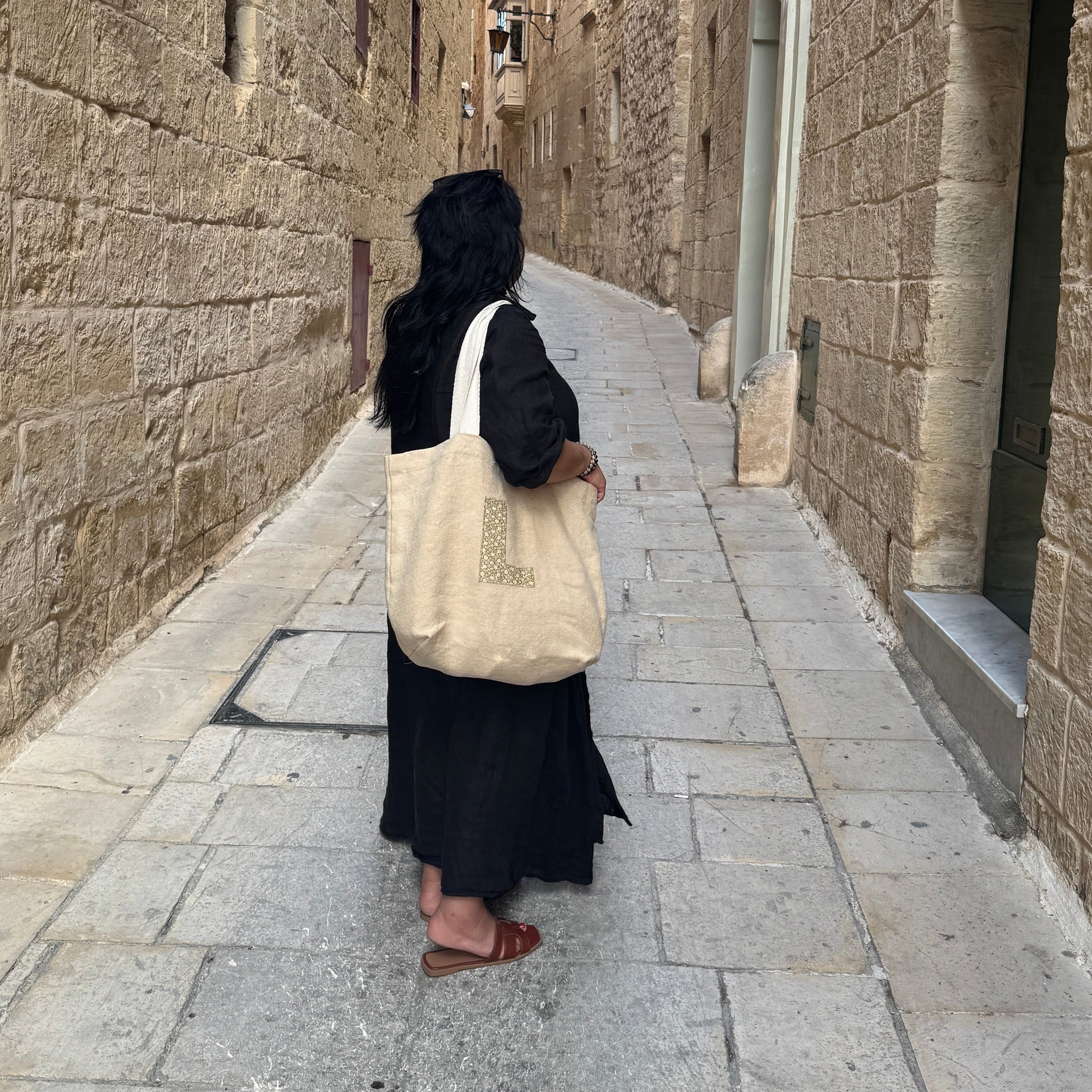 Woman in a long black dress looking down a long narrow street on her shoulder is a beige tote bag showing the letter L on the front 