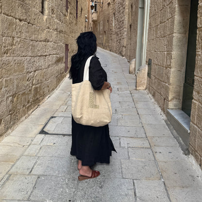 Woman in a long black dress looking down a long narrow street on her shoulder is a beige tote bag showing the letter L on the front 