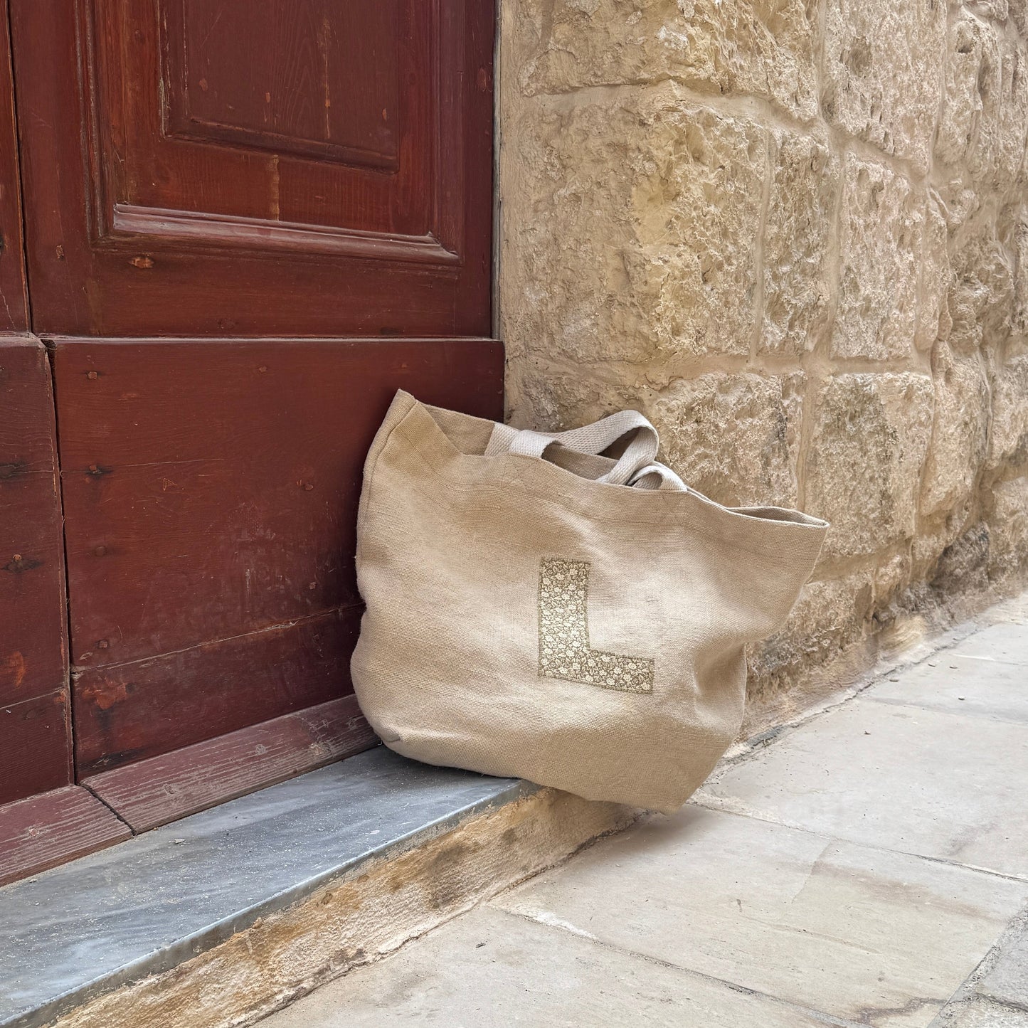 Beige bag with a large green floral letter L on the front leaning against a stone wall next to a door