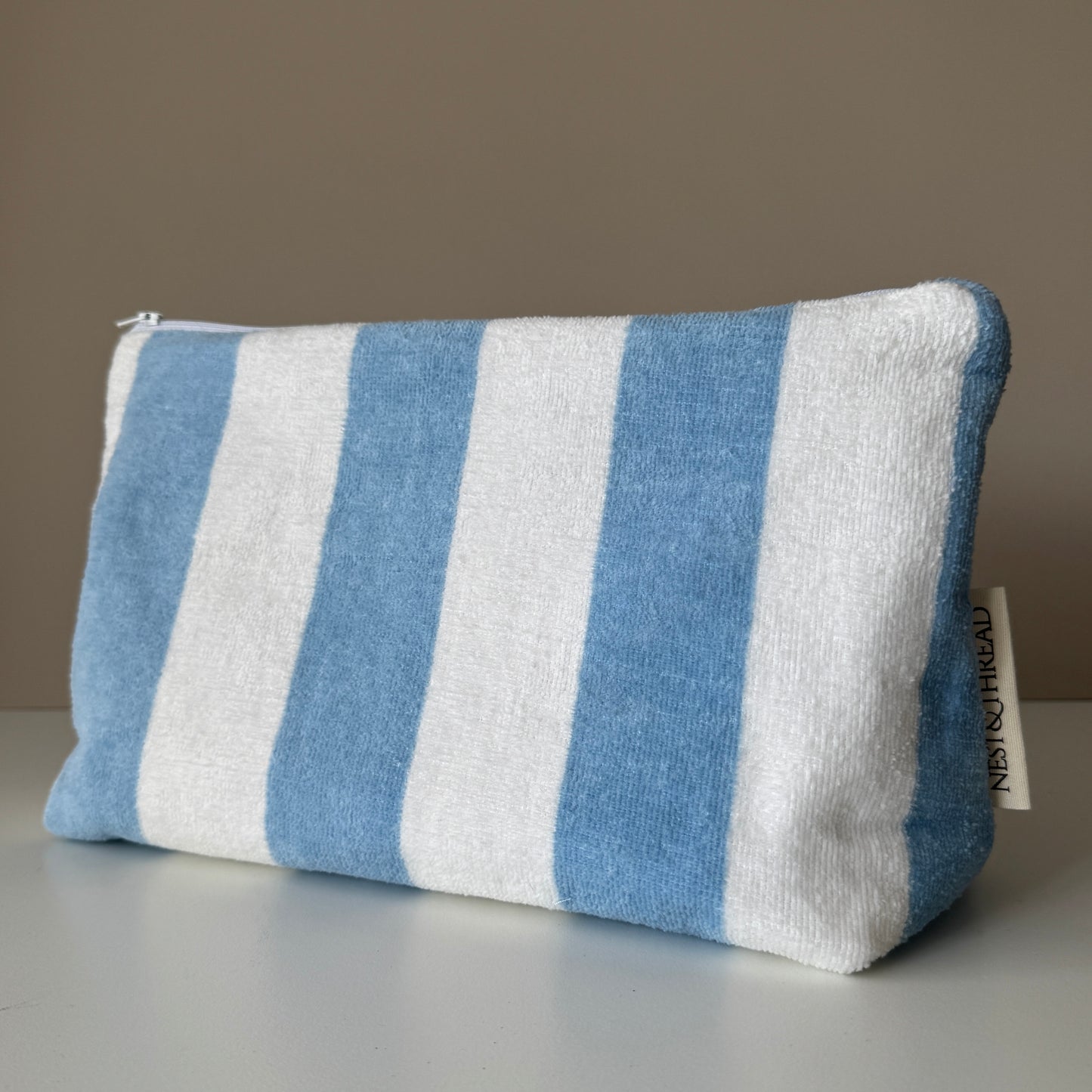 A rectangular zippered pouch made of towelling fabric with vertical blue and white stripes, shown on a neutral gray surface against a taupe background.