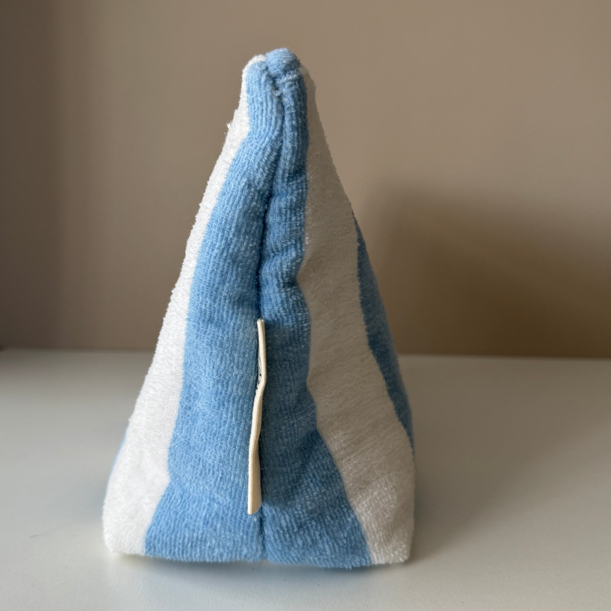 The side view of a fabric pouch or organiser made from light blue and white striped terry cloth, standing upright on a white surface against a beige background.