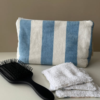 A striped blue and white towel pouch sits on a tabletop with a black hairbrush in front and reusable white face wipes to the right, against a plain beige wall.
