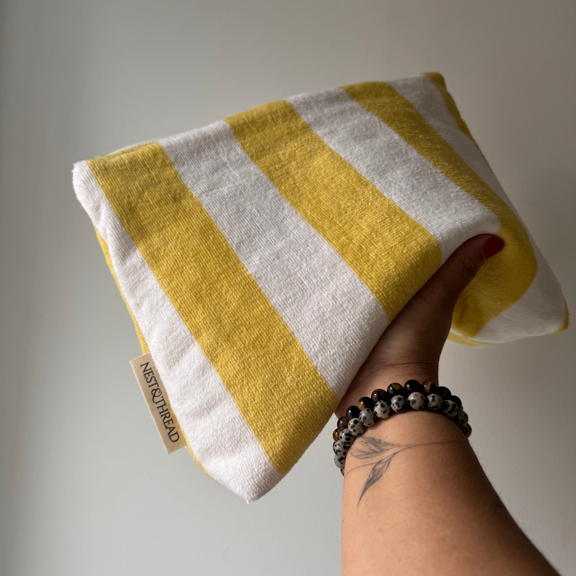 A hand holding a towelling cosmetic or makeup pouch with alternating yellow and off-white horizontal stripes. A small fabric tag on the edge reads 'NEST AND THREAD'