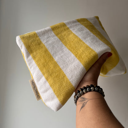 A hand holding a towelling cosmetic or makeup pouch with alternating yellow and off-white horizontal stripes. A small fabric tag on the edge reads 'NEST AND THREAD'