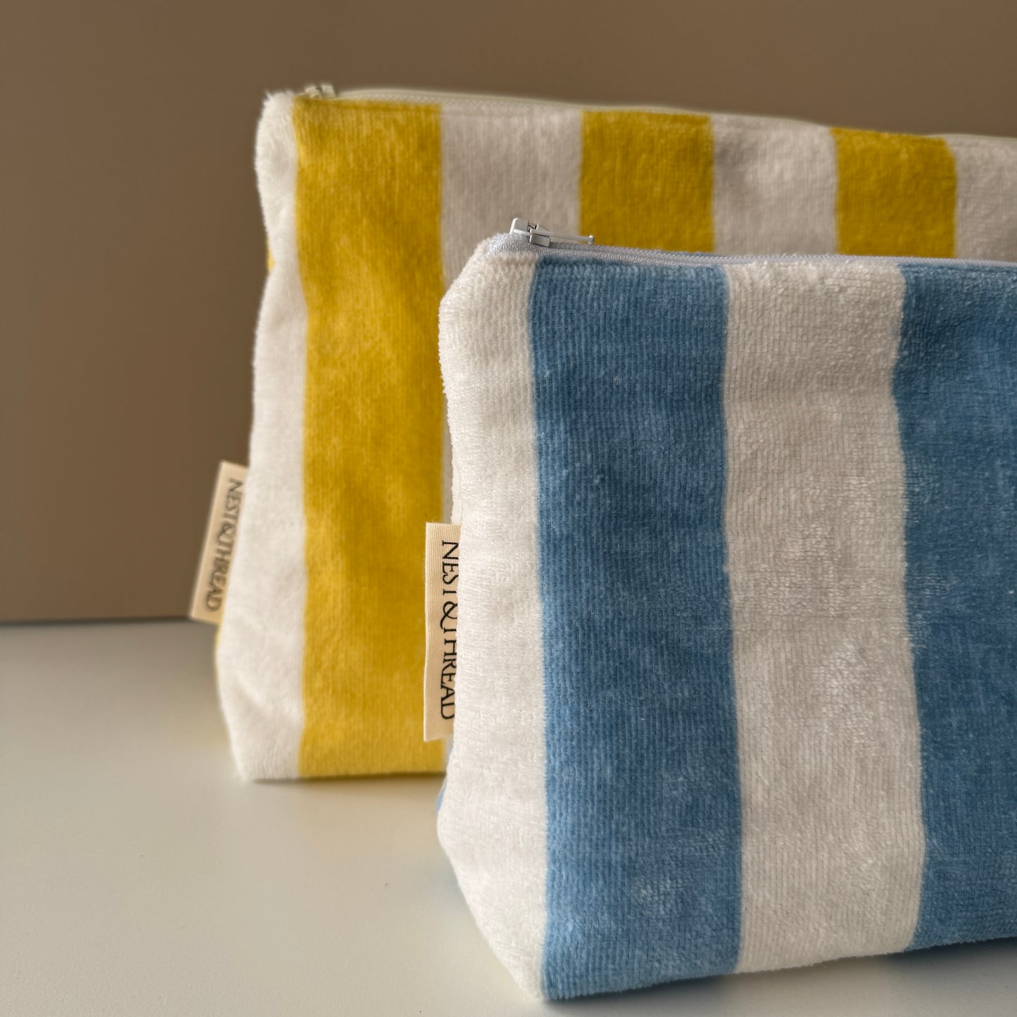 Two striped fabric zip pouches on a light surface against a warm beige background. The front pouch is white with blue and white vertical stripes, and the rear pouch is white with yellow  vertical stripes.