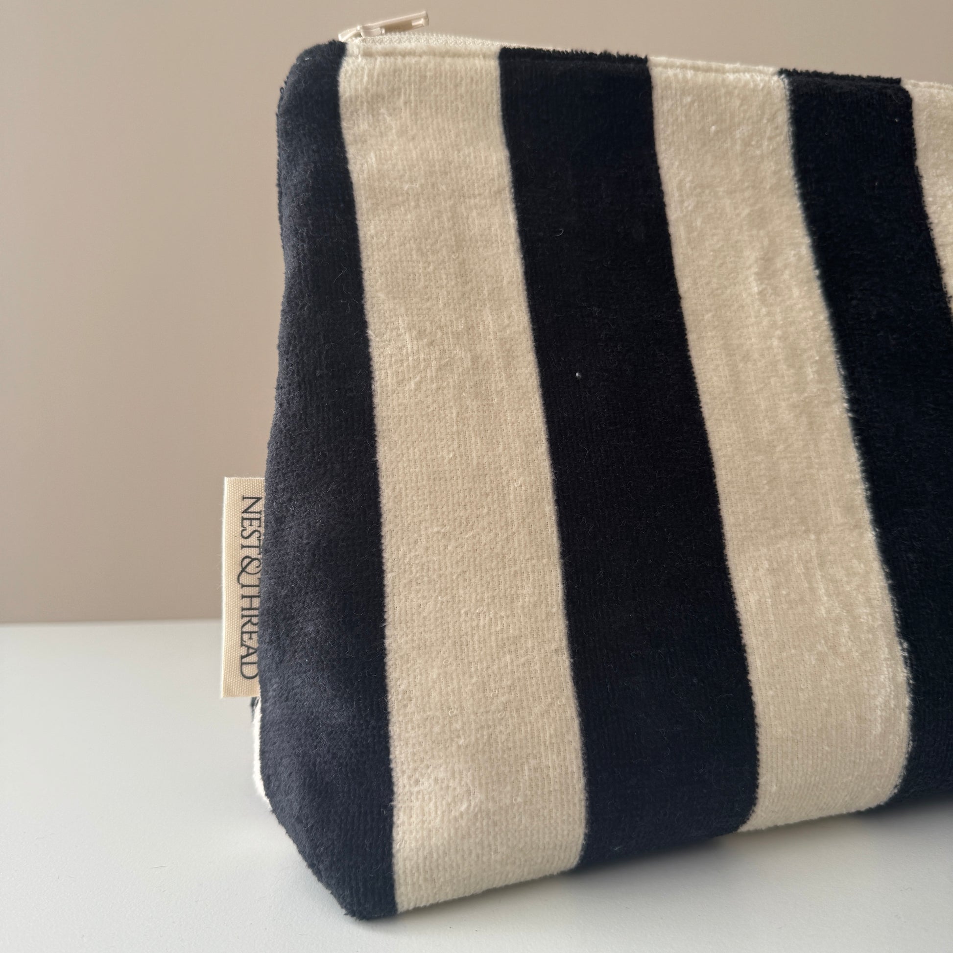 A striped fabric pouch with alternating black and cream vertical stripes, featuring a small fabric tag on the side that reads 'NEST AND THREAD'.