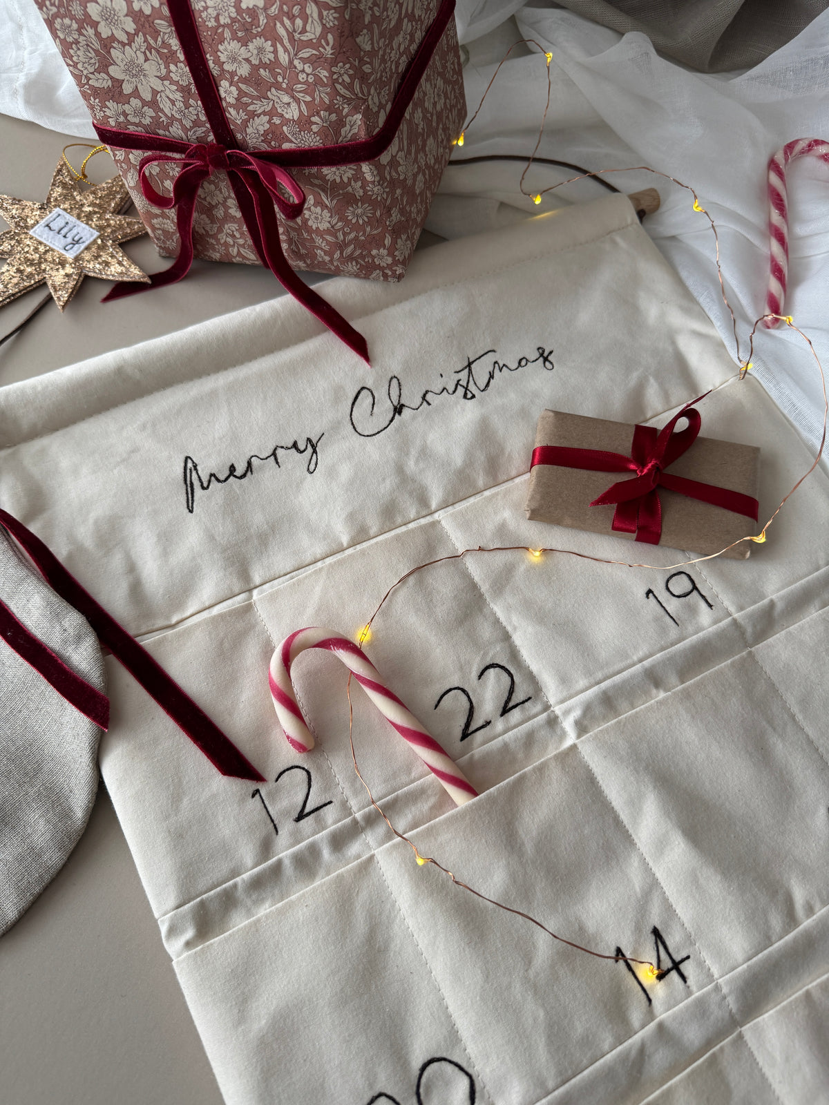 A Christmas advent calendar made from cream-colored quilted fabric, featuring stitched compartments labeled with numbers and the script text 'Merry Christmas'. The calendar is styled with red velvet ribbons, a small wrapped gift, candy canes, fairy lights, and a patterned gift box on a neutral background.