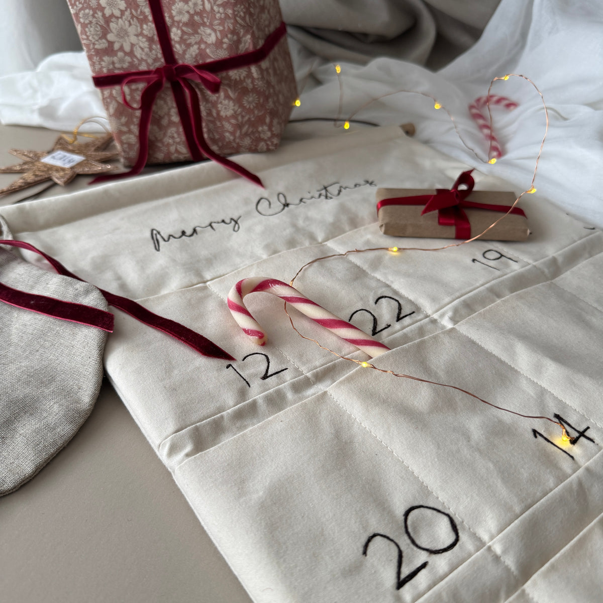 A Christmas advent calendar made from cream canvas with stitched numbered compartments, embroidered 'Merry Christmas' text, and dark red ribbon ties. Candy canes, fairy lights, wrapped gifts, and soft fabric props are arranged around it on a light surface.